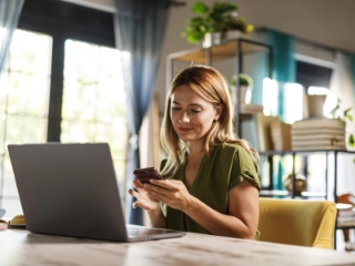 Frau sitzt zu Hause am Tisch mit offenem Laptop und Smartphone in der Hand